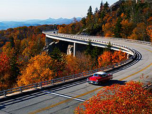 The Linn Cove Viaduct, a red car with a white top is on a road surrounded by autumn leaved trees