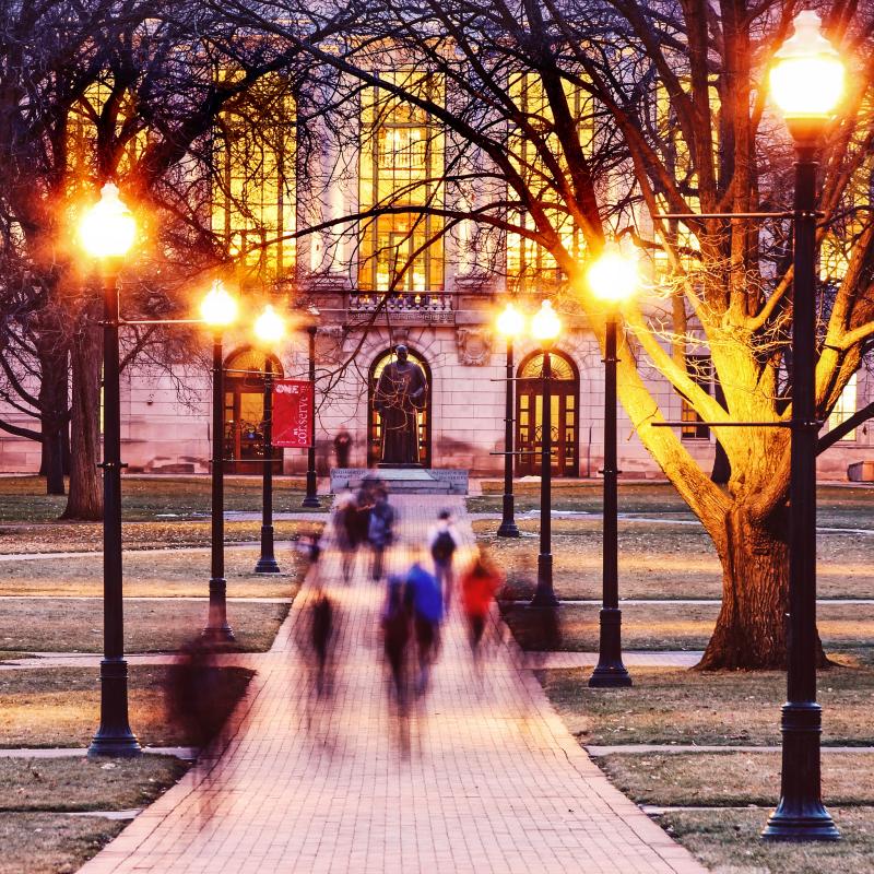 Thompson Library at night. Students walking up the sidewalk toward the building.