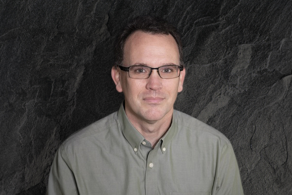 Man with dark hair and glasses in front of a dark stone wall