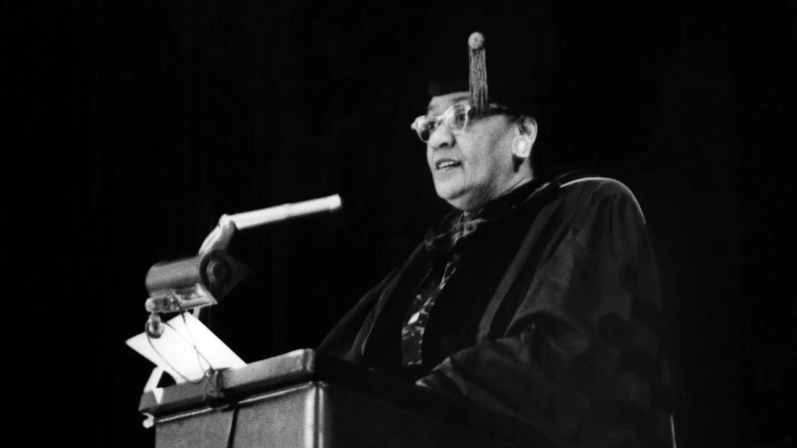 Black and white photo of a woman in graduation gown speaking at a podium