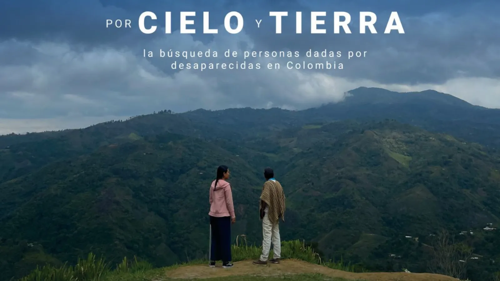 Two people standing with their backs turned facing a forested mountain on a cloudy day