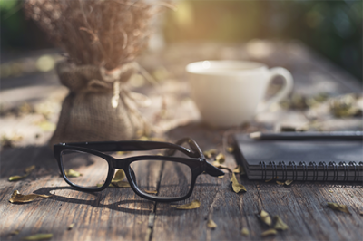 black eye glasses on a wooden table, with a white coffee cup and a notebook and pen, and a bunch of dried flowers in a burlap bag