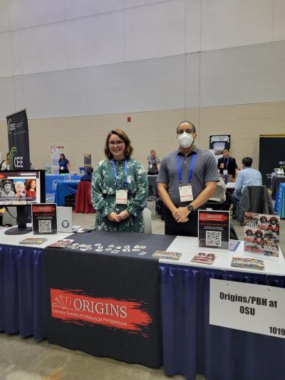 Kristin Osborne and Paul McAllister standing behind a table with postcards, stickers, signage and a computer monitor on it.