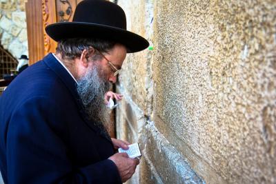 Jewish man, in front of a cement wall, reading from a prayer book.