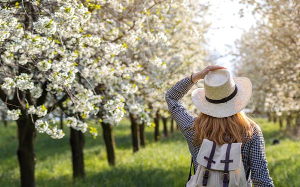 a woman wearing a backpack with her hand on her hat with white flowering trees in front of her