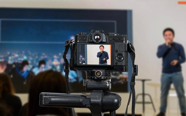 camera filming a man standing in front of a group with a microphone