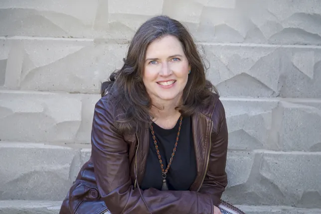 Smiling woman in a leather coat sitting in front of a stone wall