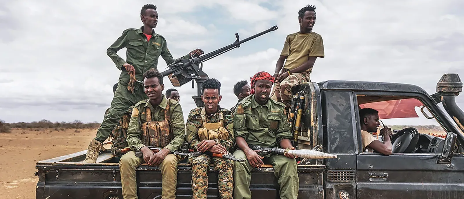 Armed fighters in military uniforms ride in the back of a black pickup truck fitted with a mounted heavy machine gun, traveling across a dry, open landscape