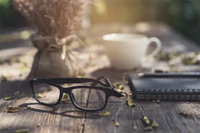 black eye glasses on a wooden table, with a white coffee cup and a notebook and pen, and a bunch of dried flowers in a burlap bag