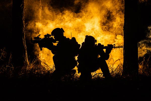two soldiers with guns raised crouching down with fire in background behind them