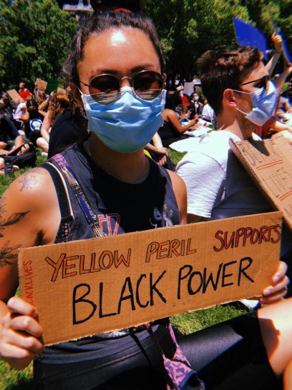 Hyun-Joo holds a sign that states "Yellow Peril Supports Black Power" at an outdoor rally in Columbus. She is wearing sunglasses and a face mask. 