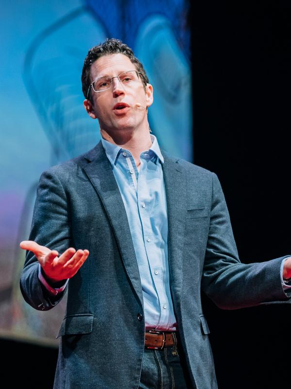 Christopher McKnight Nichols giving a TED talk, looking at the camera, hands held out above the waist palms up