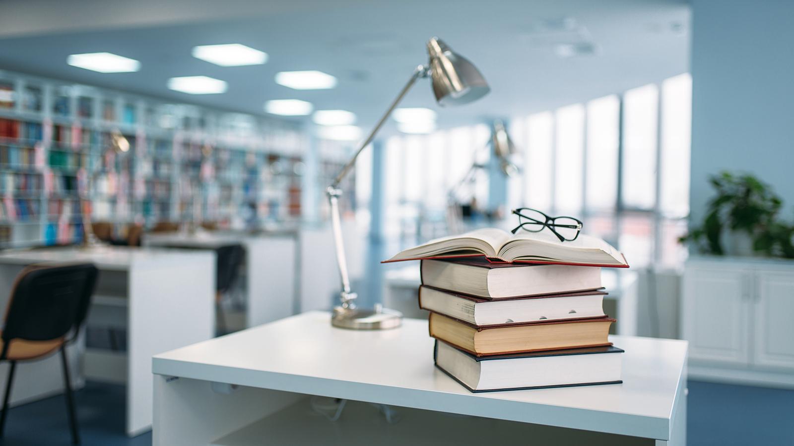 books and a pair of eye glasses on a table with a lamp in a library